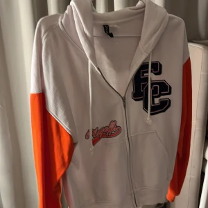 Vit och orange hoodie med dragkedja - Säljer en cool vit hoodie med orangea ärmar och dragkedja. Den har tryck med bokstäver på bröstet och en justerbar huva med snören. Perfekt för en avslappnad stil.