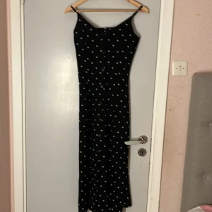 Svart prickig klänning - Snygg svart klänning med vita prickar och smala axelband. Perfekt för en stilren look. Klänningen har en lång passform som ger en elegant siluett.