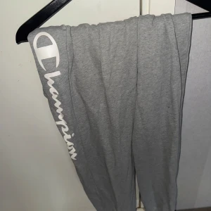 Grå mjukisbyxor från Champion - Säljer ett par grå mjukisbyxor från Champion med logga längs benet. Byxorna har en bekväm passform och är perfekta för en avslappnad stil. De är tillverkade i mjukt material och har en elastisk midja för extra komfort.