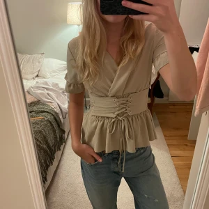 Beige omlottopp med snörning - Säljer en stilren beige omlottopp med snörning i midjan och volangdetaljer. Toppen har korta ärmar och en smickrande passform man kan ha åt båda håll! 