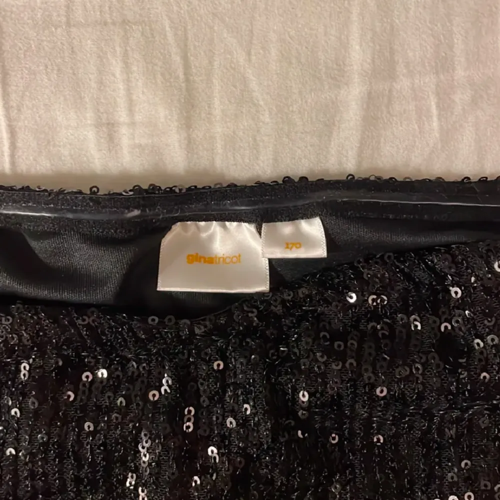 Snygg svart axelbandslös klänning med glittriga detaljer. Perfekt för fest eller nyår! Den har en smickrande tight passform upptill och en volangkjol som ger en fin siluett. Materialet är stretchigt och bekvämt, vilket gör den lätt att bära hela kvällen.. Mekot.