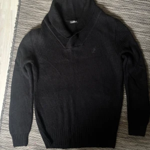 Svart stickad tröja från J.Lindeberg - Säljer en stilren svart stickad tröja från J.Lindeberg. Tröjan har en hög krage och långa ärmar, perfekt för kyligare dagar. Den ribbade designen ger en klassisk look. Passar bra till både jeans och chinos.