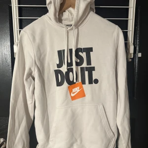 Vit hoodie från Nike - Säljer en stilren vit hoodie från Nike med det klassiska 'Just Do It' trycket i svart på framsidan. Hoodien har en bekväm passform med långa ärmar och en praktisk ficka framtill. Perfekt för en avslappnad stil.