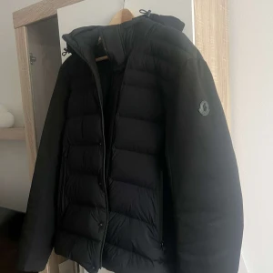 moncler doudoune elastique jacka  - Säljer en stilren svart dunjacka från Moncler. Jackan har en quiltad design med dragkedja och knappar framtill. Den är långärmad och har en huva för extra skydd mot kyla. Perfekt för kalla dagar!