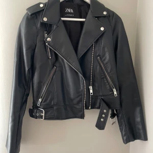 Svart skinnjacka från Zara - Snygg svart skinnjacka från Zara med dragkedjor och bälte. Jackan har en klassisk bikerstil med långa ärmar och silverfärgade detaljer. 