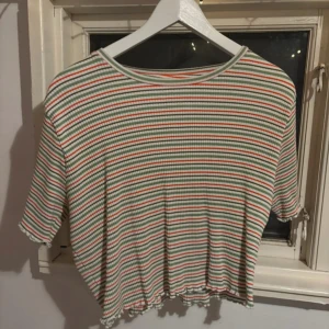 Randig ribbad topp - Randig ribbad topp med korta ärmar. Toppen har en mix av gröna, vita och röda ränder och en vågig kant vid nederdelen. Perfekt för en avslappnad stil. Croppad modell, ej använd. Strl XL men skulle snarare beskriva den som M-L beroende på hur man vill att den ska sitta.