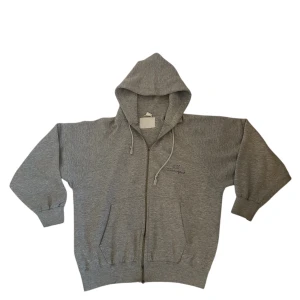 Grå Champion zip-up hoodie - Säljer en klassisk grå hoodie från Champion med dragkedja och huva. Tröjan har långa ärmar och ribbade muddar vid ärmslut och nederkant. Perfekt för en oversized stil. Som du ser har en del av dragkedjan lossnat dock. Annars har den inga fler defekter än det.