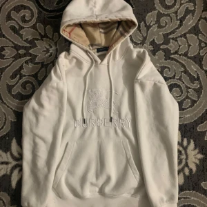 Vit hoodie från Burberry - Säljer en stilren vit hoodie från Burberry med broderad logga och ikoniskt mönstrad huva. Perfekt för en avslappnad och trendig stil, tröjan är helt ny och är i storlek S 