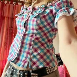 Rutig blus med spetsdetaljer - Charmig blus i blått och rosa rutmönster med vita spetsdetaljer. Blusen har korta ärmar och söta knappar med hjärtan framtill. Den är dekorerad med spännen vid axlarna. Går att bära både off och on shoulder. Storlek S.