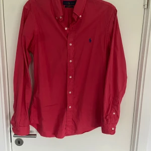 Röd skjorta från Ralph Lauren - Snygg röd skjorta från Ralph Lauren i slim fit. Tillverkad i lätt twillmaterial med klassisk krage och knappar framtill. Den har långa ärmar och en broderad logga på bröstet. Perfekt för en stilren look.