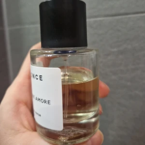 Parfymflaska med svart lock - Elegant parfymflaska med en minimalistisk design. Flaskan är genomskinlig med en vit etikett och har ett svart lock. Innehållet är en ljusgul vätska som ger en lyxig känsla.