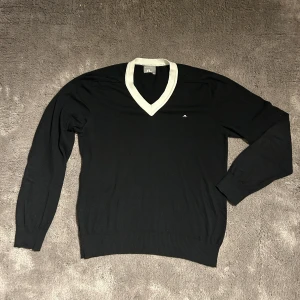 Svart tröja med vit v-ringning från J.Lindeberg - Snygg svart tröja från J.Lindeberg med vit v-ringning. Tillverkad i 100% bomull, perfekt för en stilren look. Den har långa ärmar och en liten logga på bröstet.