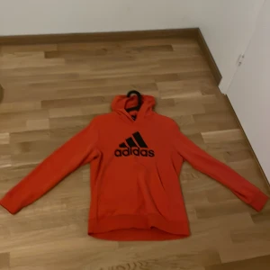 Orange hoodie från Adidas - Säljer en snygg orange hoodie från Adidas med deras klassiska logga i svart på framsidan. Tröjan har långa ärmar och en bekväm huva. Perfekt för en sportig look eller avslappnade dagar.