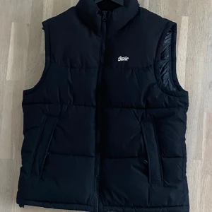 Svart dunväst från Pull&Bear - Snygg svart dunväst från Pull&Bear med dragkedja framtill. Perfekt för lager-på-lager under kyliga dagar. Västen har en stilren design med diskret logga på bröstet.