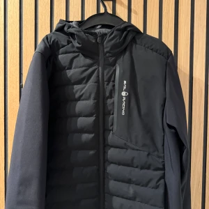Svart jacka från Sail Racing - Snygg svart jacka från Sail Racing med huva och dragkedja. Jackan har en cool logga på bröstet och en patch på ärmen. Perfekt för kyliga dagar med sin quiltade design. En ficka vid sidan om bröstet och två bra fickor på sidorna där nere! Storlek S och den är bra i storleken! Skicket är bra men den är använd så man kan väl se att den är använd. 