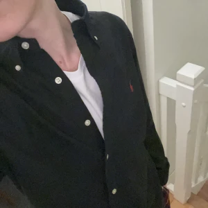Svart skjorta från Polo Ralph Lauren - Snygg svart skjorta från Polo Ralph Lauren med röd broderad logga på bröstet. Skjortan har långa ärmar och knappar framtill. Perfekt för en stilren look.