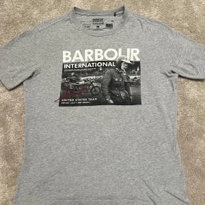 Barbour T-Shirt - Snygg grå Barbour T-shirt med tryck. Perfekt skick, endast använd 1-2 gånger. Inga defekter. Köptes på Ströms i somras men har ej kommit till användning. Storlek M. Köparen står för fraktens bekostnad. 