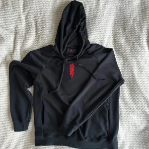 Svart Zion hoodie från Jordan - Säljer en snygg svart hoodie från Jordan med röd grafik på bröstet och ryggen. Perfekt för både vardag och träning. Den har en bekväm passform och är tillverkad i mjukt material. Huvan har dragsko för justering. Passar perfekt till höst och vinter! 