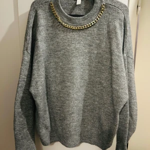 Grå stickad tröja med guldkedja - Säljer en superfin grå stickad tröja från H&M. Den har en cool guldkedja runt halsen som ger en extra touch. Tröjan är långärmad och perfekt för höst och vinter. Den är i storlek M och är riktigt mysig och varm. Perfekt för att hålla stilen när det blir kallt ute!