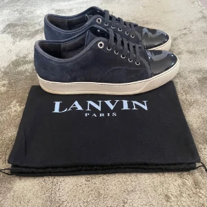Mörkblå sneakers från Lanvin - Säljer ett par snygga mörkblå sneakers från Lanvin. De har en stilren design med en kombination av mocka och lackdetaljer på tån. Skorna har en vit sula och klassisk snörning. Perfekta för både vardag och fest!