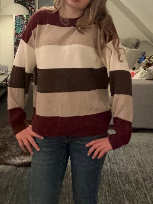 Randig stickad tröja i jordnära toner - Stickad tröja med breda ränder i vinrött, beige, brunt och vitt. Tröjan har rund halsringning och långa ärmar. Perfekt för dig som gillar en avslappnad och trendig stil. Materialet känns mjukt och bekvämt. Alrig använd, tog bara på för passform