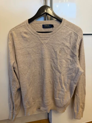 Beige tröja från Polo Ralph Lauren - Snygg beige tröja från Polo Ralph Lauren med rund halsringning och diskret broderad logga på bröstet. Tröjan har långa ärmar och ribbade muddar vid ärmslut och nederkant. Perfekt för en clean och klassisk stil.