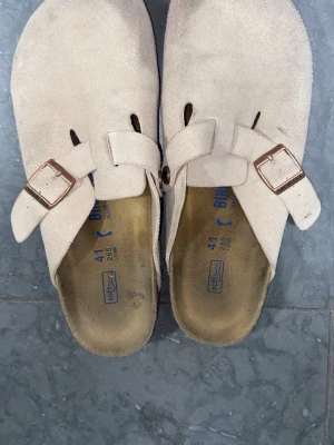 Beige mockatofflor med spänne - Säljer ett par klassiska beige tofflor från Birkenstock i mjuk mocka. De har justerbara remmar med metallspännen och en bekväm fotbädd i kork. Perfekta för dig som gillar stilrena och tidlösa skor med skön passform.
