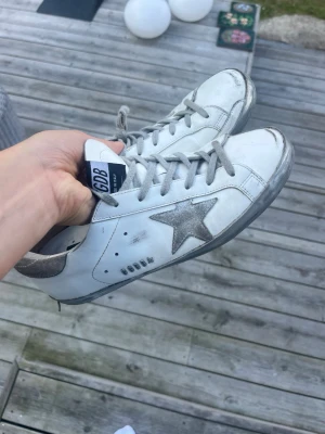 Vita golden goose sneakers - Snygga vita sneakers från Golden Goose med grå/silvrig stjärna på sidan och grå/silvriga detaljer. Är köpta från vestiaire collective för 4000 där de har blivit kollade för äkthet, originalpriset tror jag ligger mellan 5000-6500 för denna modell, om ni har några frågor svarar jag gärna på allt!❤️ De är inte superslitna men som ni kan se är sulan lite lite uppriven på ena hälen👍 kan tänka mig att gå ner i pris vid snabb affär!