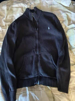 Mörkblå zip hoodie från Polo Ralph Lauren - Mörkblå hoodie med dragkedja från Polo Ralph Lauren. Klassisk design med ribbade muddar, två framfickor och broderad logga på bröstet. Tillverkad i mjukt stickat material som ger en avslappnad och stilren look. 1800 ny. Passar från 160-175 cm 