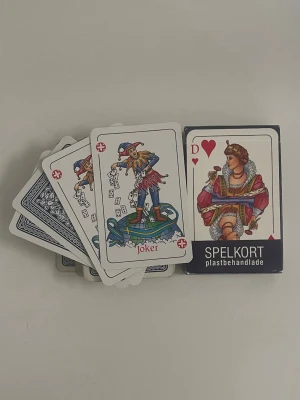 Kortlek plastbehandlade spelkort - En klassisk kortlek med plastbehandlade spelkort. Korten har traditionella motiv, inklusive joker och dam i hjärter. Baksidan har ett blåvitt mönster och framsidan färgglada illustrationer. Perfekt för kortspel med vänner eller familj.