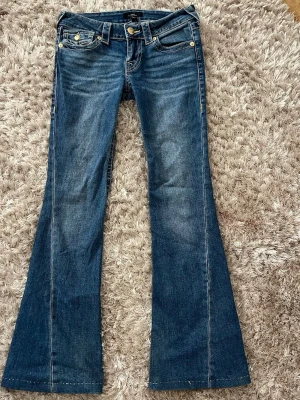 True religion joey bootcut jeans - Snygga blå bootcut jeans med låg midja och klassiska fem fickor. Jeansen har en lätt tvättad look och utsvängda ben. Tillverkade i denim med kontrastsömmar och metallknappar. Perfekta för en trendig och avslappnad stil. Handsydd på benen som är ca 73 cm i inerbenssömn.