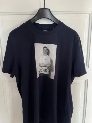 Mörkblå t-shirt med tryck - Snygg mörkblå t-shirt med ett stort svartvitt tryck framtill som visar en person och en signatur. T-shirten har rund hals och korta ärmar. Perfekt för dig som gillar stilrena plagg med en twist.