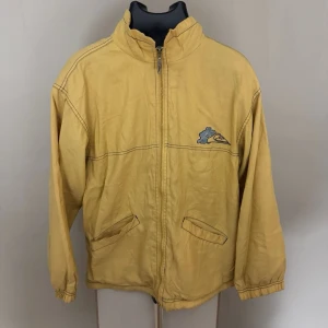 Gul vindjacka från Quiksilver - Gul vindjacka från Quiksilver med dragkedja framtill, två snedställda fickor och broderad logga på bröstet samt stor logga med klöver på ryggen. Junior 14 år. 0390