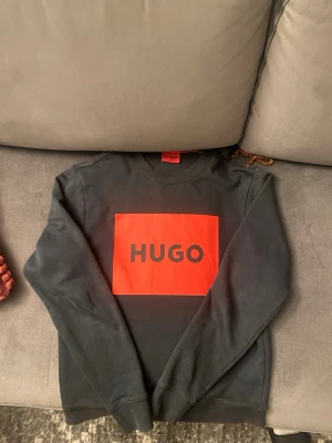 Svart sweatshirt från HUGO - Cool svart sweatshirt från HUGO med stor röd logga på bröstet. Tröjan har rund halsringning och långa ärmar. Perfekt för dig som gillar streetwear och vill ha en enkel men snygg look.