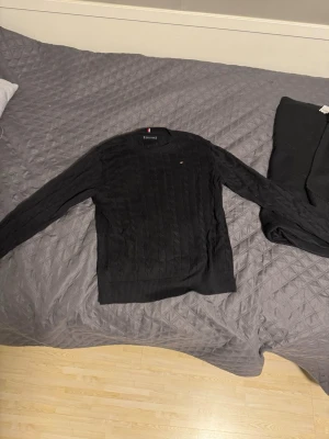 Svart kabelstickad tröja Tommy Hilfiger - Svart kabelstickad tröja från Tommy Hilfiger med diskret logga på bröstet. Tröjan har rund halsringning, långa ärmar och ribbade muddar. Perfekt för en stilren och klassisk look. Sorry för dålig bild på taggen, hade brottom. Hesitera ej om att skriva för frågor/prisdiskutiom