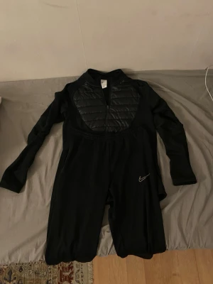 Svart overall från Nike med vaddering - Svart långärmad overall från Nike med dragkedja framtill och vadderad panel över bröstet. Overallen har en sportig design, elastisk midja och Nike-logga på benet. Perfekt för träning utomhus under kallare dagar.