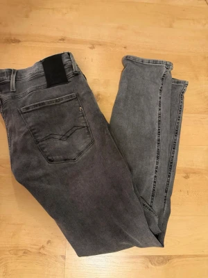 Replay jeans  - Säljer ett par grå slim fit jeans med svarta detaljer vid linningen och snygga sömmar på bakfickan. Jeansen har klassisk femficksdesign och är tillverkade i ett mjukt denimtyg med lite stretch för skön passform.