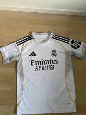 Real Madrid matchtröja Bellingham - Vit Real Madrid matchtröja från Adidas med svarta detaljer och gula linjer. Tryck med 'Emirates Fly Better' på framsidan och 'Bellingham 5' på ryggen. Klubbmärke och Adidas-logga på bröstet. Perfekt för fotbollsfans!