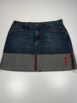 Phard Kort jeanskjol med röd söm - Snygg kort jeanskjol i blå denim med kontrasterande grå nederkant och röda sömmar. Kjolen har klassiska fickor fram och bak samt bälteshällor. Unik detalj med röd brodyr på nederdelen framtill. 0085