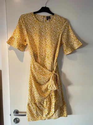 Gul blommig omlottklänning Vero Moda - Superfin gul kortklänning från Vero Moda med vitt blommönster. Klänningen har omlottdetalj med knyt i midjan, volangkant nertill och korta ärmar. Perfekt för sommarens alla tillfällen!
