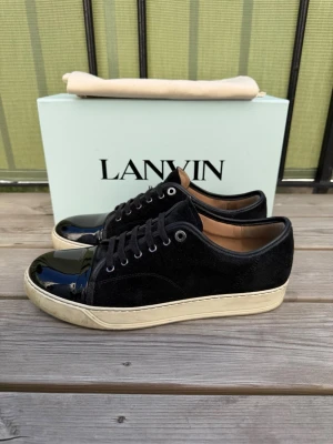 Svarta Lanvin sneakers med lackdetalj - Snygga svarta sneakers från Lanvin med ovandel i mocka och glansig lackad tå. Skorna har vita sulor och svarta skosnören. Perfekt för dig som gillar stilrena och exklusiva sneakers med en twist. Lite defekt på ena tån som ses på bilderna. Bara skor kommer med