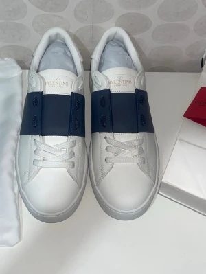 Valentino vita sneakers med blå detalj - Snygga vita sneakers från Valentino Garavani med bred mörkblå läderdetalj över snörningen. Klassisk låg modell med rund tå och vita skosnören. Tillverkade i slätt läder för en clean och exklusiv look. Aldrig använda, pris kan diskuteras🤗