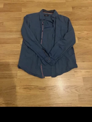 Blå långärmad skjorta med knappar - Säljer en stilren blå skjorta med långa ärmar och klassisk krage. Skjortan har knappar framtill och en bröstficka. Passar perfekt till jeans eller chinos för en avslappnad look.