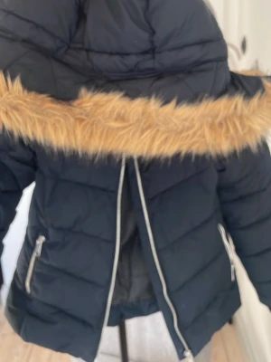 Svart dunjacka barn vinter  - Snygg svart dunjacka med stor huva och beige fuskpälsdetalj runt kanten. Jackan har två dragkedjeförsedda fickor och quiltad design som ger en modern look. Perfekt för kalla vinterdagar. Till för barn storlek 146!🤗
