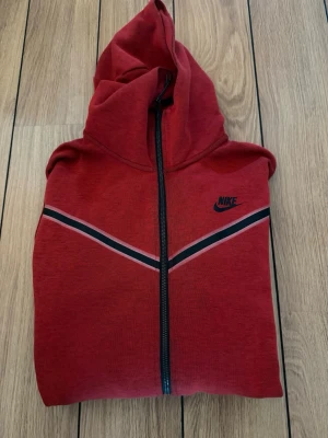 Röd Nike hoodie med dragkedja - Snygg röd hoodie från Nike med hel dragkedja framtill och klassisk huva. Tröjan har svarta och vita ränder i V-formad design över bröstet samt Nike-logga på vänster sida. Perfekt för en sportig och avslappnad stil.