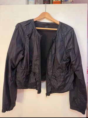 Svart kroppad jacka från Lindex - Snygg svart vindjacka med dragkedja framtill och dragsko i midjan för justerbar passform och en mer snatched look. Jackan har långa ärmar, rundad halsringning och två fickor framtill. Tillverkad i ett lätt och glansigt material som passar perfekt för blåsiga dagar.