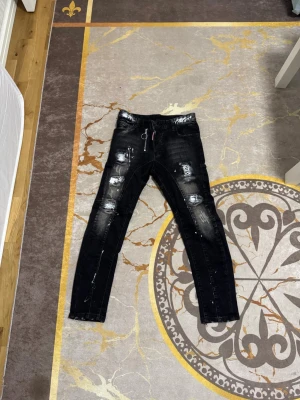 Svarta Dsquared2 jeans med detaljer  - Säljer ett par svarta skinny jeans med slitningar, vita färgstänk och dragkedjedetalj framtill. Jeansen har flera hål och patchade partier för en trendig, street-inspirerad look. Materialet är troligtvis bomull med stretch för skön passform.