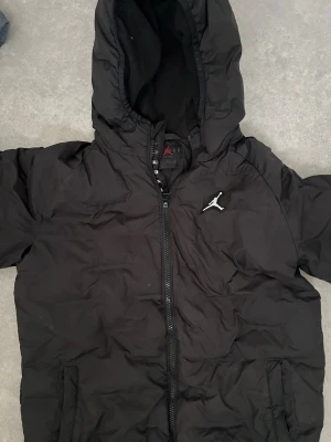 Svart dunjacka med huva från Jordan - Snygg svart dunjacka från Jordan med huva och dragkedja framtill. Jackan har den klassiska Jumpman-loggan på bröstet och två sidofickor. Perfekt för kalla vinterdagar och ger en sportig look.