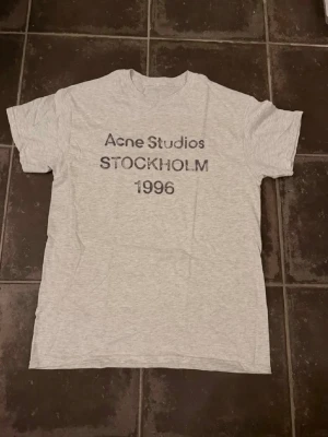 Grå Acne Studios t-shirt Stockholm - Säljer en ljusgrå t-shirt från Acne Studios med tryck 'Acne Studios STOCKHOLM 1996' på bröstet. Klassisk rund hals och korta ärmar. Perfekt för dig som gillar minimalistisk och stilren design. Är ca 182 och väger 66