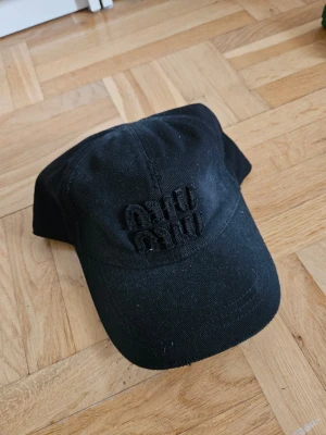 Miu Miu keps med broderad text - Säljer en svart keps i bomull med böjd skärm och broderad text framtill i samma färg som kepsen. Klassisk modell med justerbar rem bak. Perfekt för en stilren och avslappnad look.